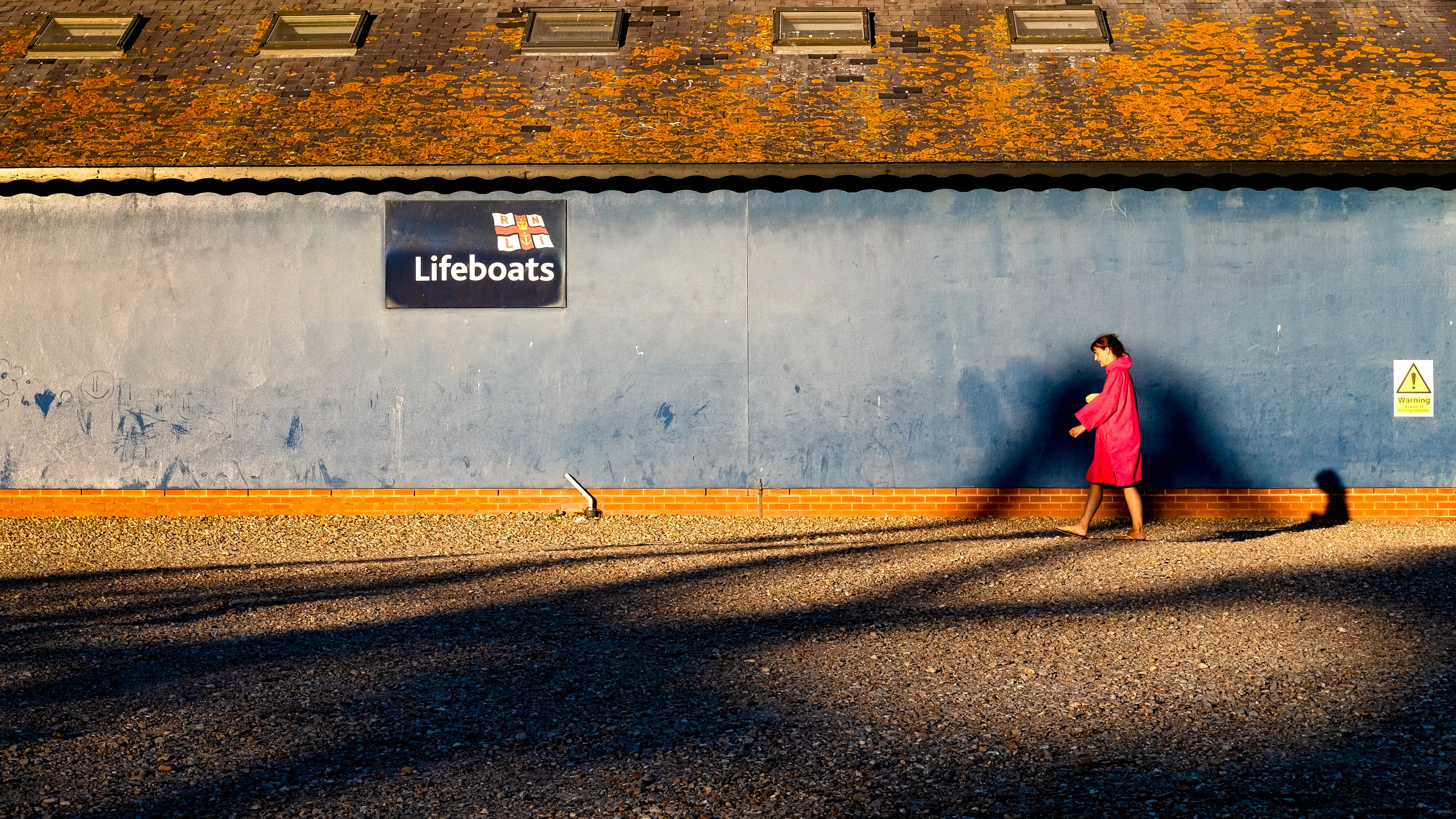 Woman in a red coat walks past an orange lit wall with a Lifeboats sign, long shadows