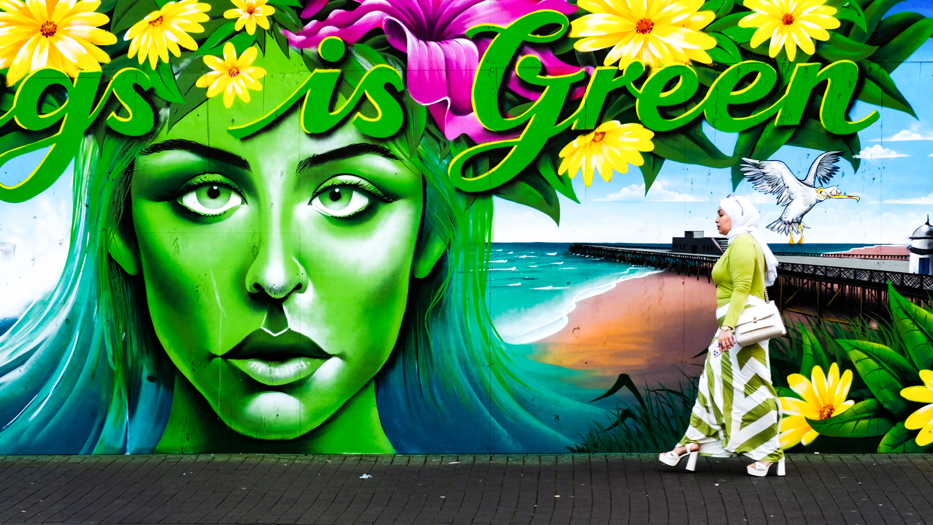 Mural of a green woman’s face with flowers, a woman walking past