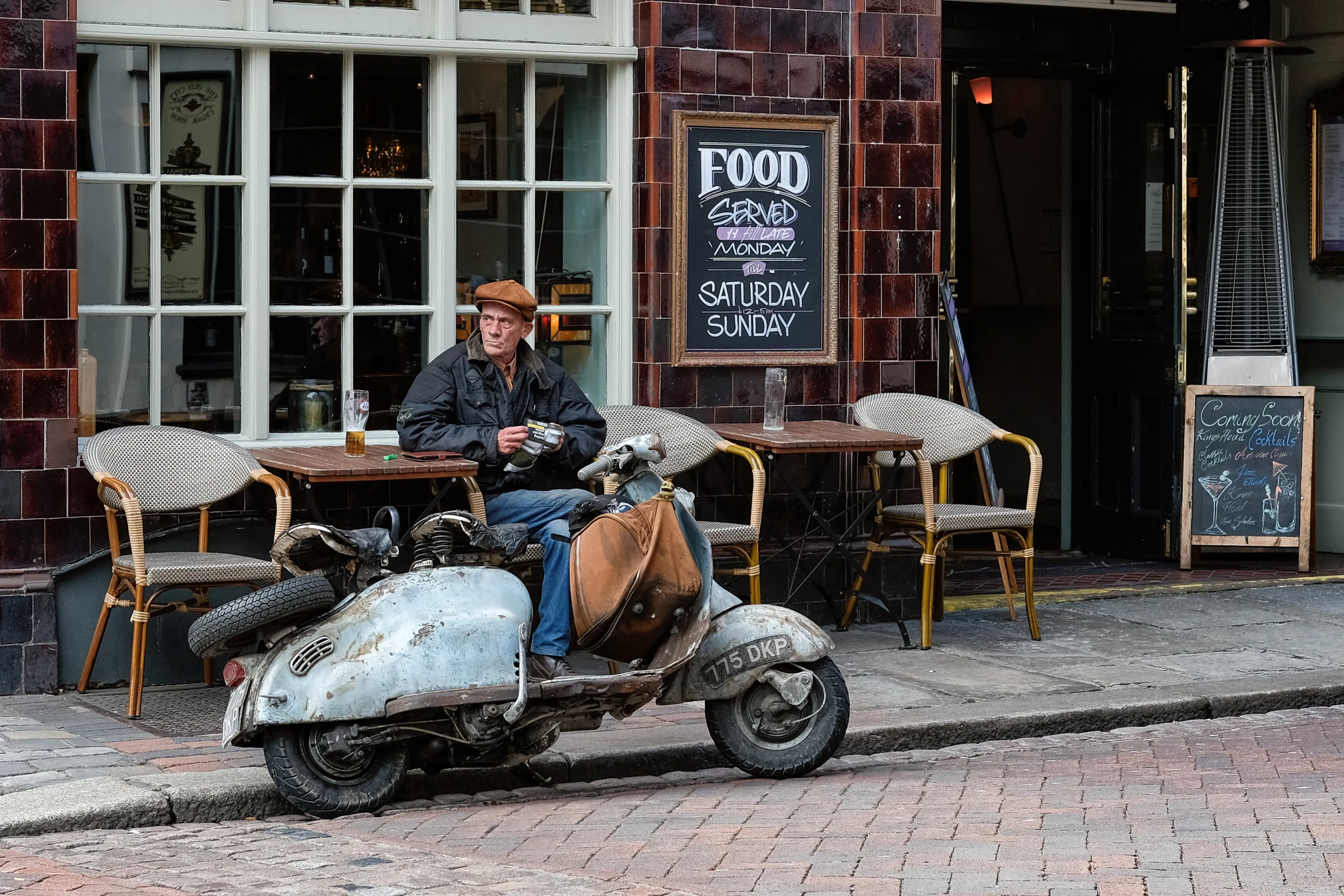 Man on a terrace beside an old scooter in front of a tiled facade