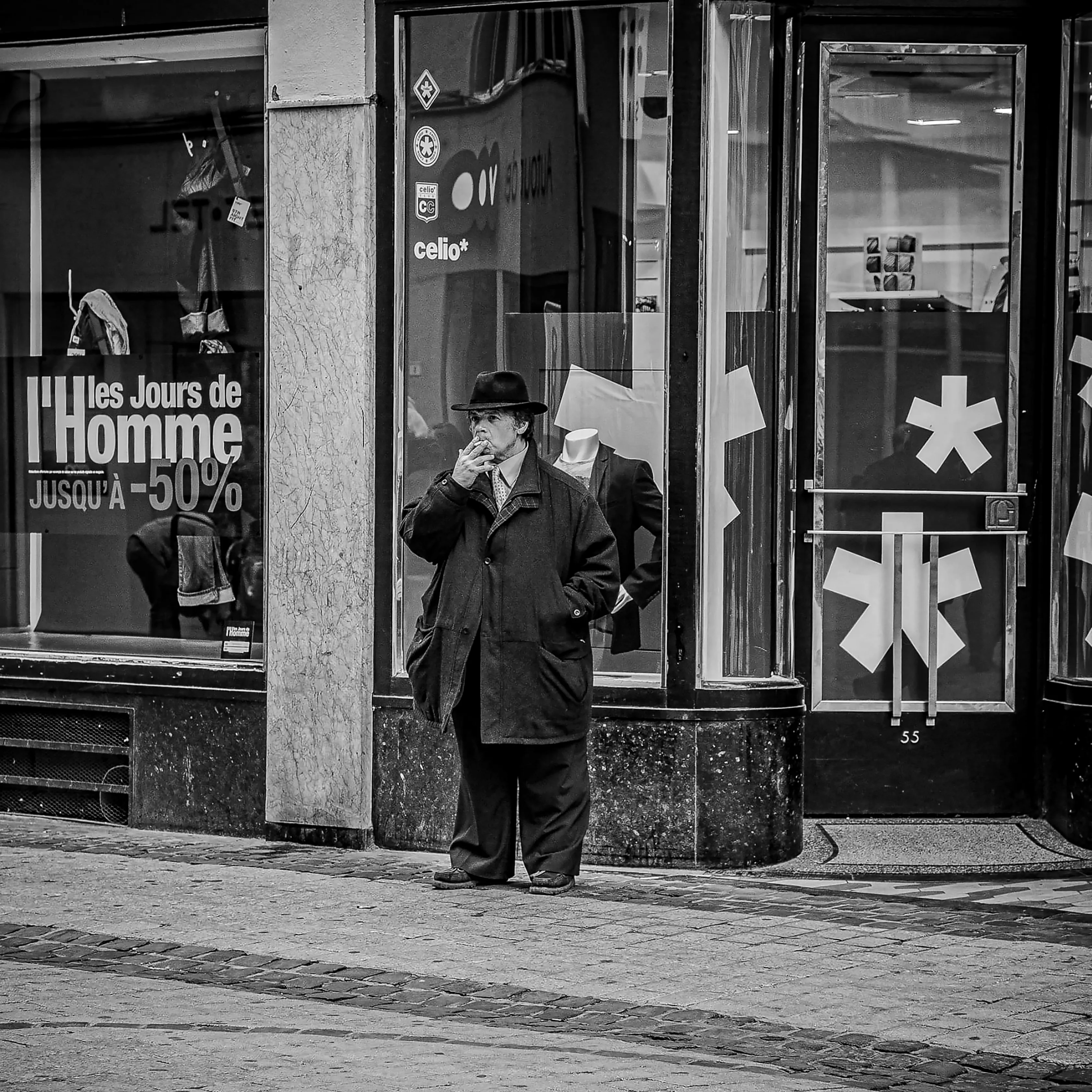 Man smoking in a long coat in front of a shop window in Liège, black and white